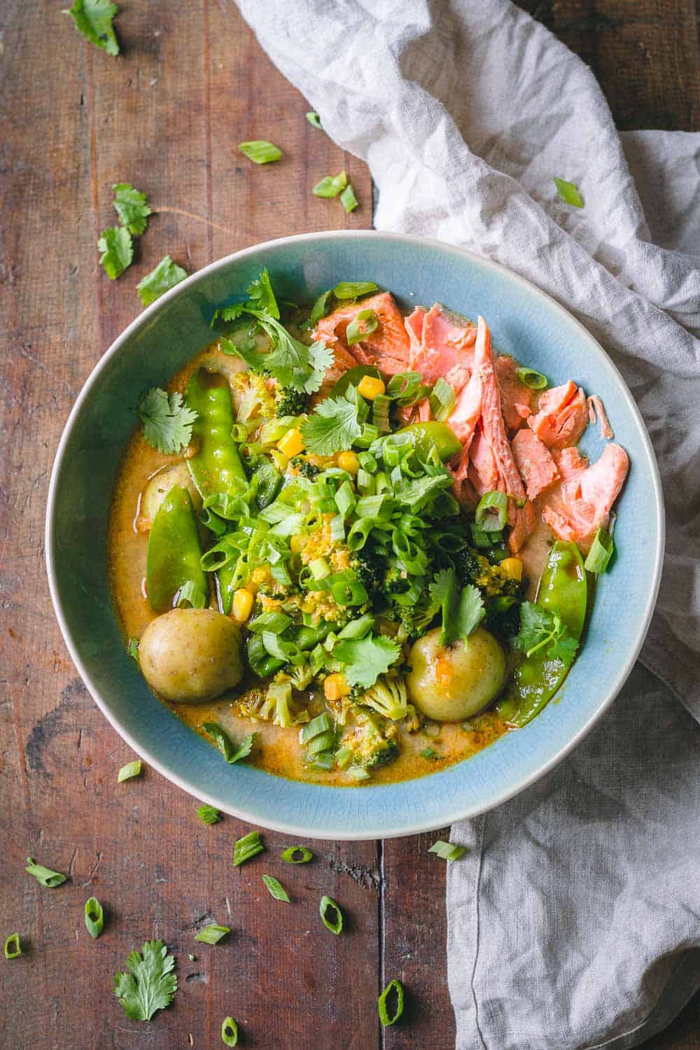 Thai Curry Soup in a blue bowl showing shredded salmon, potatoes, broccoli, and snow peas in broth on a wooden table