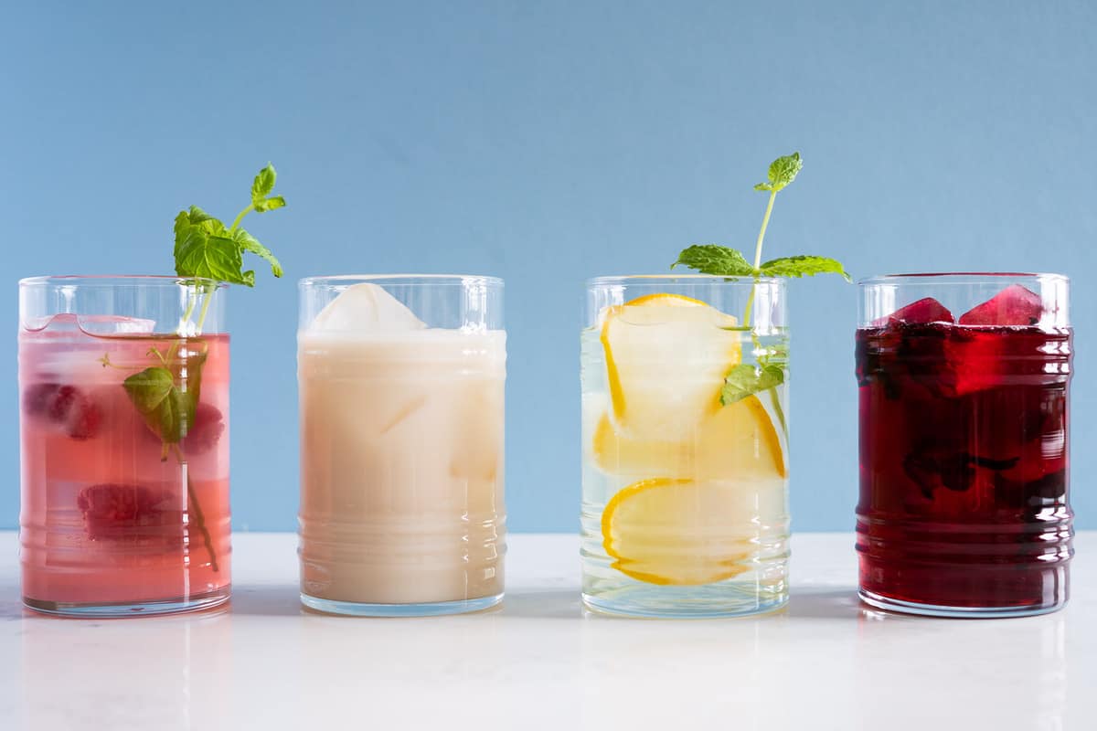 4 summer drinks - in drinking glasses - hibiscus tea, pink lemonade, agua de horchata, and lemon infused water