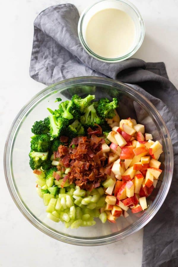 Broccoli florets, diced apple, sliced celery, and crumbled bacon in a bowl and white dressing on the side