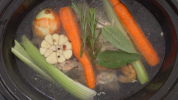 whole carrots, celery, half a bulb garlic, onion, and herbs floating in water in slow cooker
