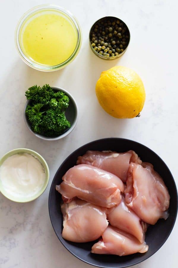 Chicken thighs in a bowl, creme fraiche in a bowl, parsley in a bowl, chicken broth in a bowl, green peppercorns in a can, and a lemon on a kitchen counter.