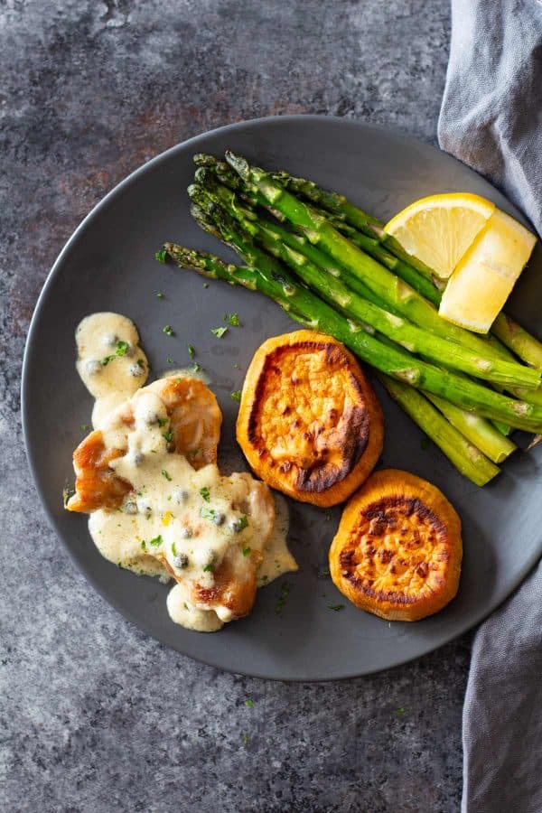 Chicken thigh with creamy peppercorn sauce on a plate served with roasted sweet potato slices and roasted asparagus.