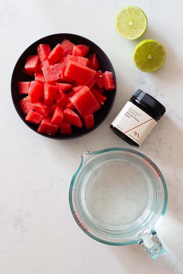 cubed watermelon in a bowl, a jug of water, a lime, and a jar of honey on a kitchen counter