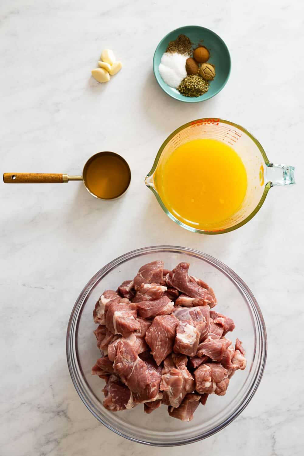 Cubed pork in a bowl, orange juice in a jug, vinegar in a measuring cup, cloves or garlic, spices on a kitchen counter.