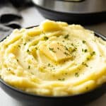 Creamy Mashed Potatoes in a bowl topped with butter and parsley, Instant Pot in background. Instant Pot Mashed Potatoes recipe