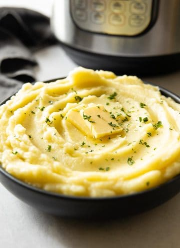 Creamy Mashed Potatoes in a bowl topped with butter and parsley, Instant Pot in background. Instant Pot Mashed Potatoes recipe
