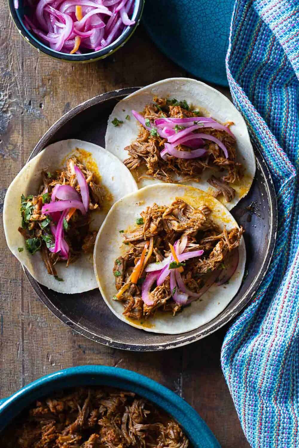 Cochinita Pibil Tacos on a wooden plate topped with pickled red onions.