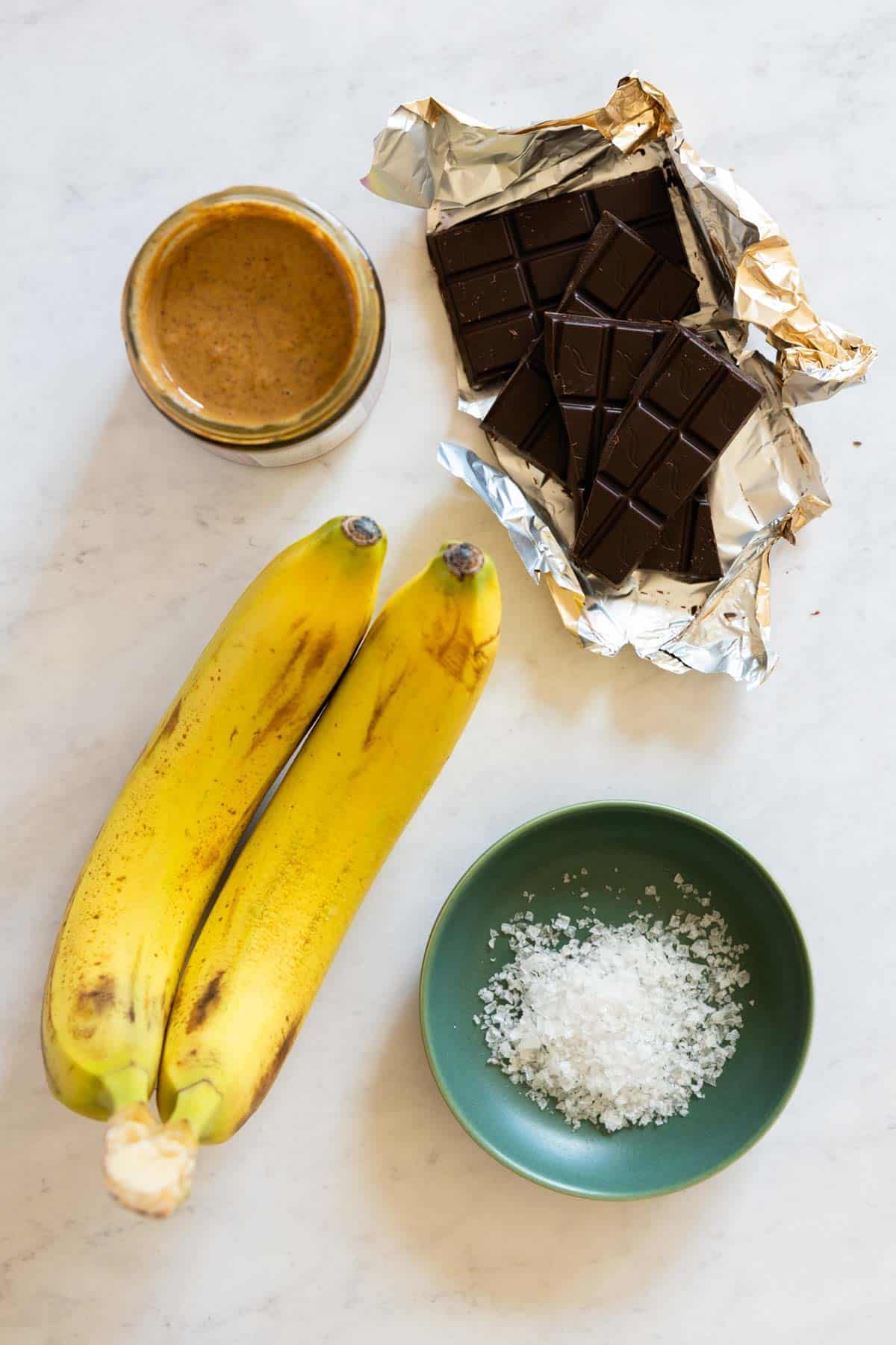 Two bananas, dark chocolate, hazelnut butter, and flaky sea salt laid out on a kitchen counter.