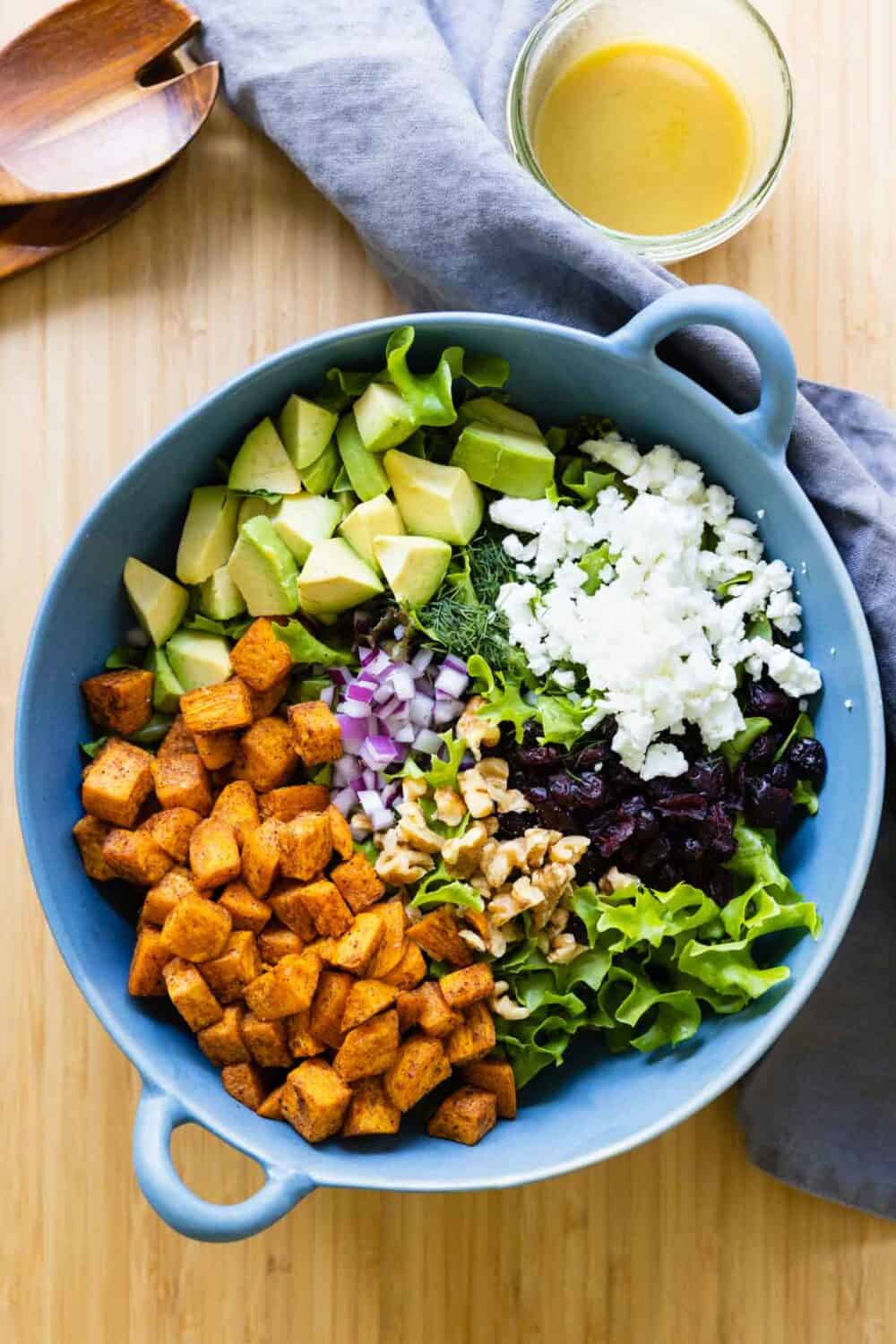 Lettuce, roasted sweet potato, avocado, onion, feta, cranberries in a blue salad bowl, dressing on the side.