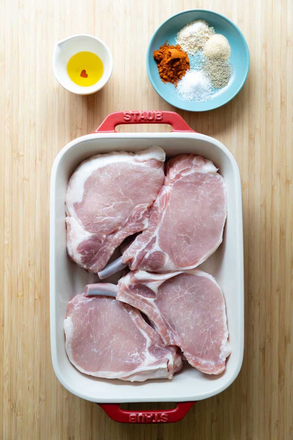 Pork chops in a baking dish, oil in a bowl, and spices on a plate laid out on a kitchen counter.