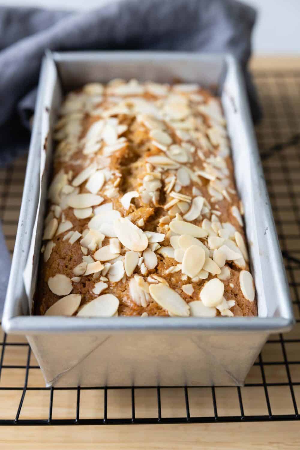 Baked apple bread with sliced almonds on top in a loaf pan on a wire rack.