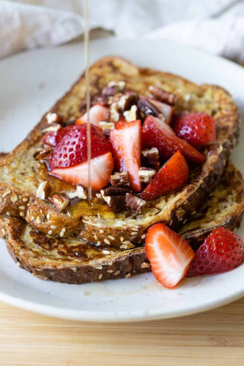 Whole-grain french toast with strawberries and nuts on top and maple syrup being drizzled on.