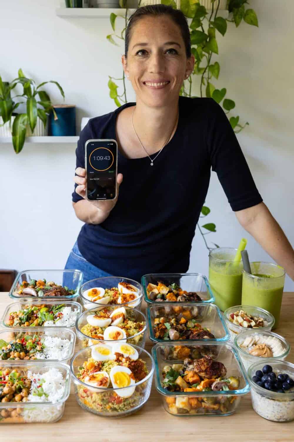 Woman holding phone that says 1 hour 9 minutes behind a counter with 15 healthy meals in glass containers.