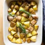 Whole garlic cloves, cut-up yellow potatoes, and rosemary in a white baking dish with red handles.