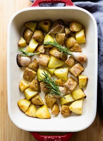 Whole garlic cloves, cut-up yellow potatoes, and rosemary in a white baking dish with red handles.