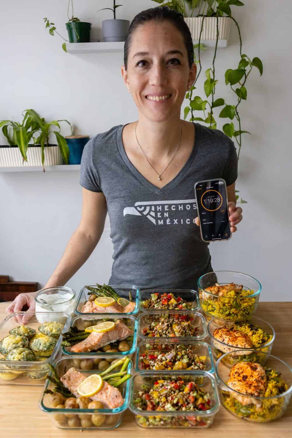 Woman standing behind 15 healthy meals in meal prep containers holding a phone reading 1:19:28.