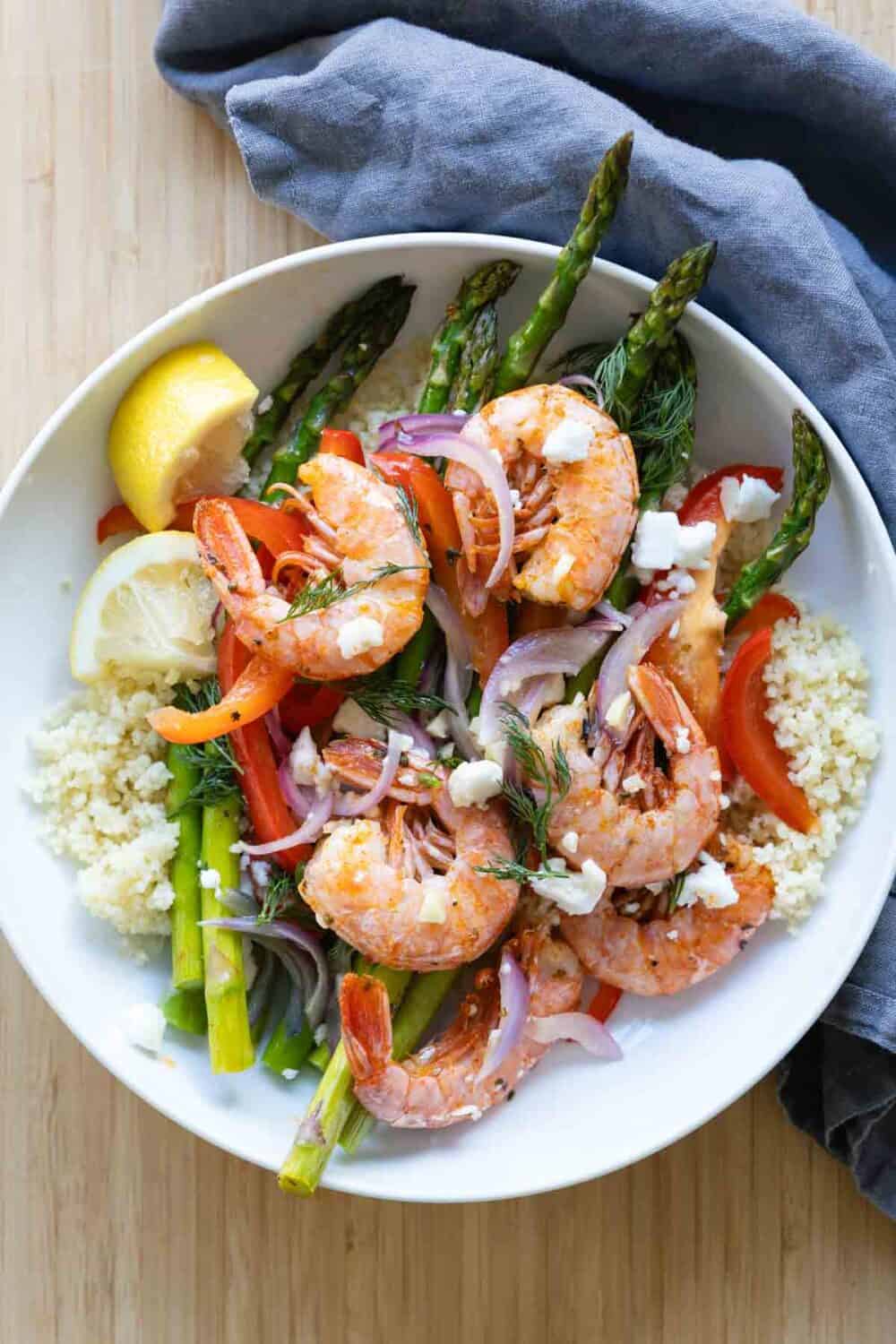 A white bowl filled with cooked couscous, asparagus, bell pepper, onion, and shrimp with lemons on the side.